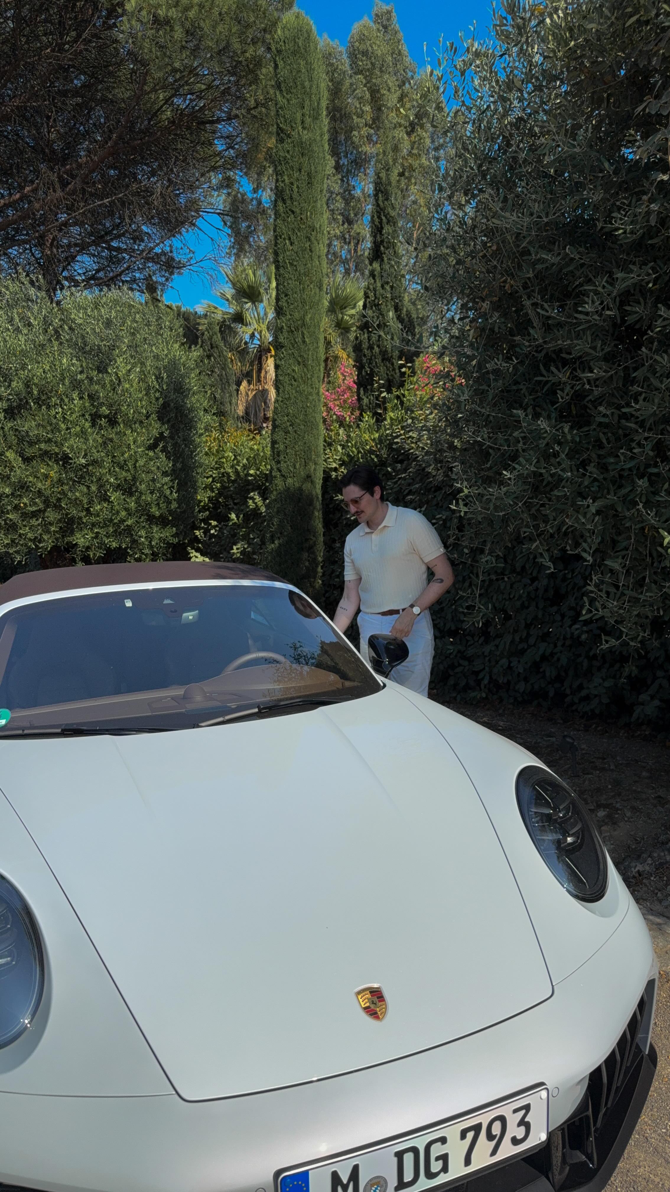 A man in sunglasses and a beige polo shirt stands near a white convertible sports car. The car is parked next to a well-landscaped area with rich green foliage. The image is shot in bright daylight.