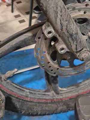 Close-up shot of a bicycle tire, brake disc, and frame component, covered with mud and dirt. The tire is partially visible with a red stripe along the edge. The disc brake has a perforated design. The background shows a floor with a repeating tile pattern. The image is shot in a low angle, emphasizing texture and details.