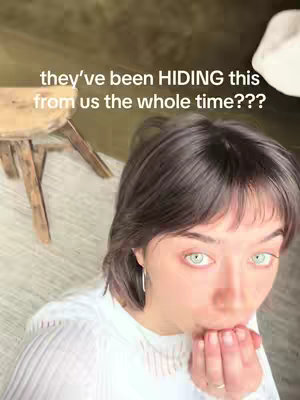 A woman with short dark hair and light eyes looks up at the camera, covering her mouth with her hand. Overlaid text reads, "they've been HIDING this from us the whole time???" The background includes a wooden stool and a light-colored floor, creating a home environment.