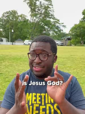 A medium shot shows a Black man with glasses, speaking directly to the camera against a backdrop of a green lawn, trees, and a cloudy sky. He is wearing a navy blue t-shirt with a white and yellow graphic that reads 'MEETING NEEDS'. The overlaid text in white reads 'is Jesus God?'