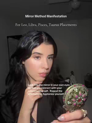 A woman with long, straight, dark hair is holding a small, ornate mirror up to her face. The image is vertically oriented, likely a selfie taken indoors. The background is softly lit, with darker tones creating potential shadows. The woman's expression seems focused. Text overlays are at the top detailing the 'Mirror Method Manifestation' and specific astrological signs. Additional text at the bottom describes key actions of the manifestation method.