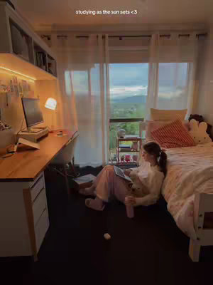 A young woman sits on the floor in her room, studying. She is leaning back against a bed, with a laptop open in front of her. The room is brightly lit, with a large window overlooking a scenic view of clouds and mountains.