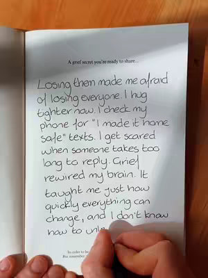 The image shows an open book on a wooden surface. The text is handwritten in black ink. There is a hand holding a pen and writing on the page. The background is blurred, with the book and the hand in focus.