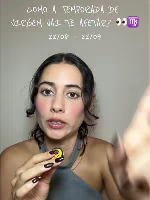 A woman is centered in the frame, with text overlayed at the top. She is holding a small, circular yellow object with a smiley face on it. She has dark hair, light skin, and is wearing a grey top. The background is a plain, light-colored wall.