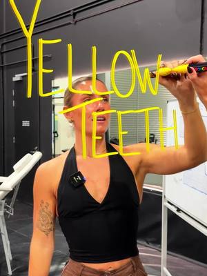 A woman is writing the words "YELLOW TEETH" in large yellow letters on a clear surface, likely a glass or mirror. She is facing the camera and smiling. The background appears to be a gym or training facility, and the lighting is bright.