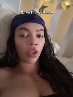 A selfie-style video cover featuring a woman lying in bed. She is wearing a blue headscarf and a necklace. The text overlay says "Planets in the 1st house". The background seems to be a white or light-colored wall.