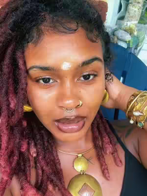 A close-up selfie of a person with dark hair styled in red-tinted faux locs. The person has dark skin, large dark eyes, gold hoop earrings, a septum nose ring, and gold bracelets on their wrist. They are wearing a black top and a gold necklace with a large pendant. They appear to be in an outdoor setting, with a hint of greenery in the background.
