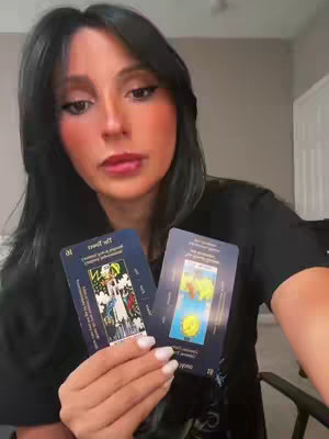 A woman with long dark hair holds two tarot cards, looking at the camera with a neutral expression. She is wearing a black top and has makeup on. The background is a neutral, light color.