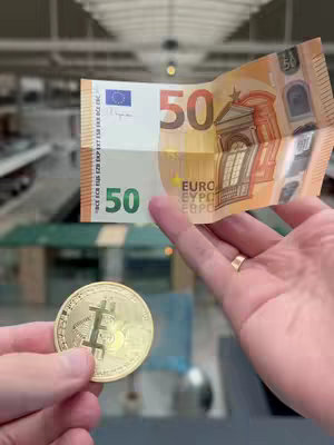 A person holding a 50 euro bill and a Bitcoin coin, with a blurred background suggesting a public space.