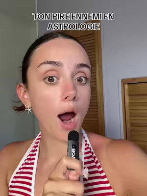 A woman with dark hair and a nose ring is centered in the image, looking directly at the camera with her mouth open, as if speaking or singing. She wears a striped halter top. Above her, overlaid in white text, reads 'TON PIRE ENNEMI EN ASTROLOGIE'. The background is a light-colored wall with a wooden door frame visible on the right side. A microphone is held close to her mouth.