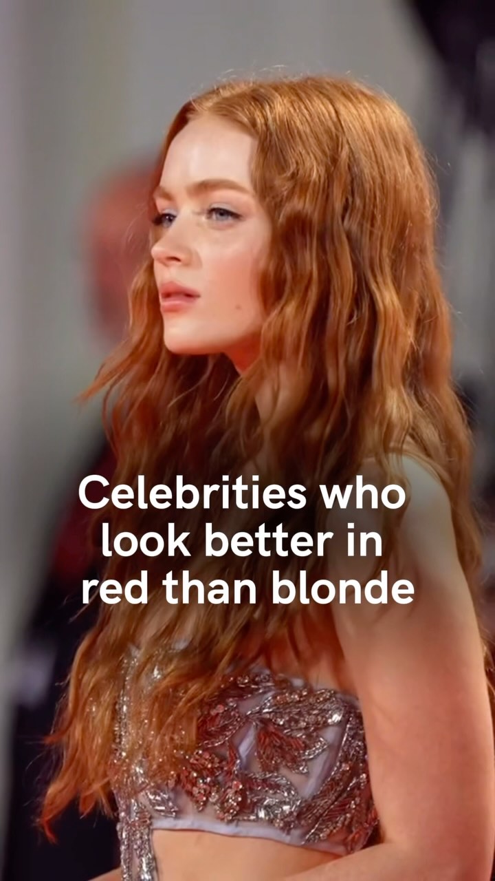 A medium shot of a woman with long, wavy red hair, looking slightly off-camera. She is wearing a formal dress with intricate detailing. Text overlayed reads, 'Celebrities who look better in red than blonde.' The background is blurred, indicating an event or photoshoot setting.