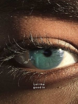 Close-up of a human eye with a light teal iris. The eye is set against a backdrop of textured skin, with prominent eyelashes and visible veins. The lighting suggests a soft, diffused source, highlighting the eye's details. The text 'Let the good in' is overlaid.