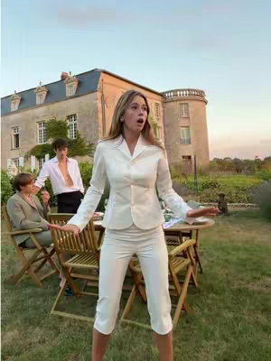 A woman in a white pantsuit stands in front of a large house and a group of people, with her arms outstretched and an expression of surprise. The scene is outdoors with a gentle blue sky.