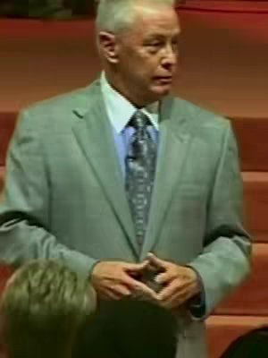 The image shows a man in a grey suit, white shirt, and patterned tie, standing in what appears to be a church or auditorium. His hands are clasped in front of him. The background features wooden panels and seating, indicating a setting for a sermon or lecture.