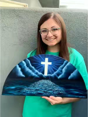 A woman with glasses holds a half-circle shaped painting, with a cross in the middle, a pathway to the cross with ocean-like waves, and a blue sky. She is smiling, wearing a green shirt and standing in front of a gray wall.