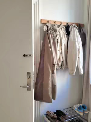 An interior shot of a hallway with a door on the left and a wall on the right. A wooden rack with five hooks holds several jackets and coats. Below, a shoe rack holds various pairs of shoes. The neutral colors of the walls and clothing create a clean, minimalist aesthetic.