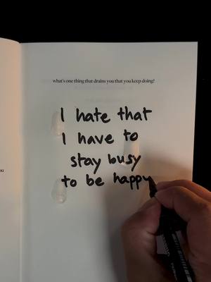 The image shows an open book with text written in black marker. The text, in a handwritten style, says, \"I hate that I have to stay busy to be happy.\" A person's hand holding a black marker is visible in the lower right, as if in the process of writing. The paper has water stains on it. The background is a dark, solid color.