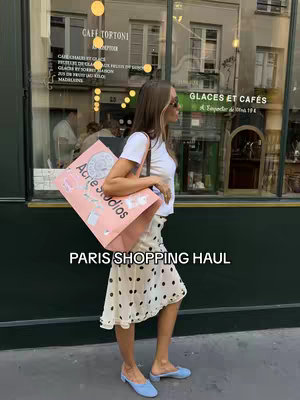 A woman stands on a street in front of a shop, showcasing a light pink shopping bag from Acne Studios. She wears a white t-shirt, a polka-dot skirt, and blue shoes. The image uses natural lighting, and the background consists of a shop window and a building facade, conveying a Parisian shopping experience.