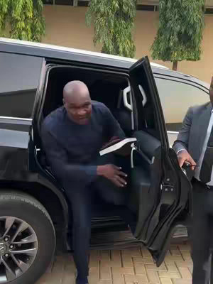 A man in a dark blue suit exits a black SUV. The door is open, and another person in a suit partially appears on the right side of the frame. Green trees are visible in the background.