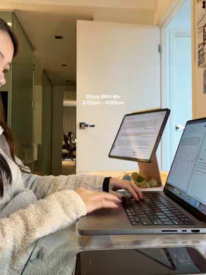 A woman sits at a desk, studying with a laptop and a tablet. The room is well-lit, with a neutral color palette dominant. A door and hallway lead to the background, suggesting depth. She is working on the laptop and has a tablet on a special stand.