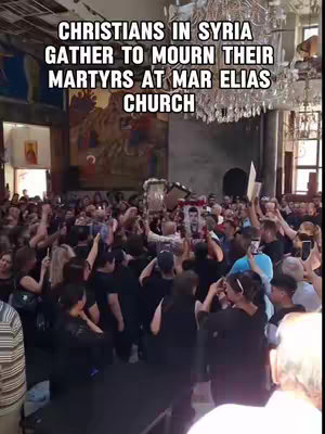A crowded interior shot of a church, filled with a large group of people. The image is dominated by a large crowd of people standing closely together in a hall with high ceilings. Many are raising their hands, and some are holding up framed photos. The focus is on capturing the emotional moment and conveying the somber atmosphere. The overall color palette is somewhat dark and subdued, dominated by blacks and grays from clothing and shadows.