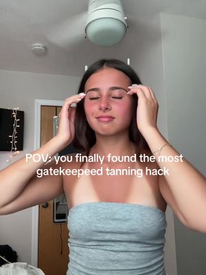 A selfie-style image of a young woman in a grey strapless top, standing in a room. Her expression is serene with closed eyes, and she is holding her hands up towards her temples. Most of her body is visible, and she has a defined tan line across her chest and shoulders. The background includes a ceiling fan, a door frame, and a small part of a wall, suggesting a bedroom setting. Text overlayed on the image indicates a tanning hack.