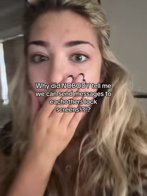 A selfie-style shot featuring a young woman covering her mouth with her hand, expressing surprise. On-screen text asks why she wasn't told about sending messages to each others' lock screens. The background is a blurred indoor setting.
