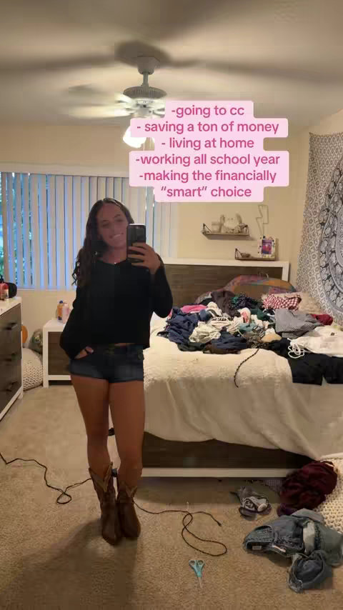 A young woman in a bedroom is taking a selfie. She is smiling and holding a phone in front of her face. On the screen, text bubbles highlight the reasons for her decision of going to community college instead of the university. The room is cluttered, with clothes and other items on the bed and a dresser.