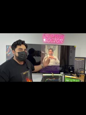 A man in a black mask and t-shirt stands in front of a room, pointing to a large screen displaying a man giving a presentation. A 'Crypto Doctor' neon sign sits above the screen. Various devices related to finance are on the table below it, including a running crypto chart.