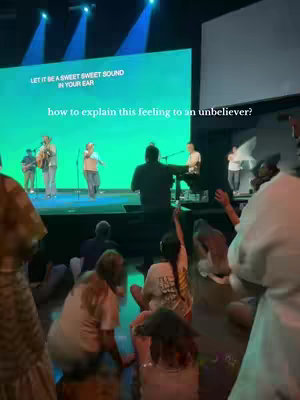A wide shot captures a worship service in a large hall. A large screen displays lyrics in green, and a band performs on a stage. The audience is seated on the floor, facing the stage, with many people raising their hands.