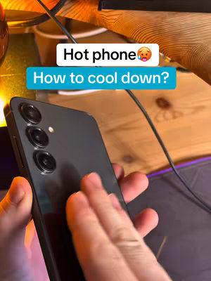 A person's hands are shown holding a black smartphone, with the screen and back of the phone facing the viewer. The background includes a wooden surface, and a cable, suggesting the phone is in an indoor environment. Text overlays 'Hot phone 🥵' is placed above it saying 'How to cool down?'