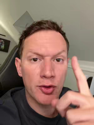 A close-up shot of a man with short, brown hair is pointing upwards with his right index finger. He has a slightly furrowed brow and is looking directly at the camera, seemingly in the middle of speaking. The background is a lightly colored room, with a black chair visible behind him. The lighting is soft and natural.
