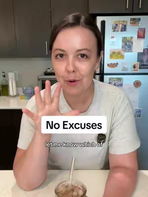 A woman is in the foreground, centered. She is speaking into the camera with her hand up near her face. Behind her is a blurred background of a kitchen with a refrigerator and dark cabinets.