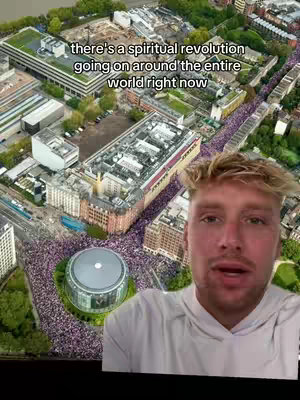 An aerial view of a large city gathering with a person in front of the video. Large buildings with people flooding the streets. The person filming is a fair-skinned man with short blond hair, wearing a white shirt. The background shows a large crowd gathered in an open space around a building.