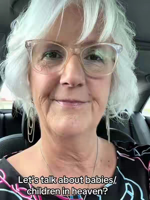A close-up shot featuring a smiling older woman with short, wavy white hair and clear-framed glasses. She is wearing a black and white patterned top. The background is a blurred view inside a car.