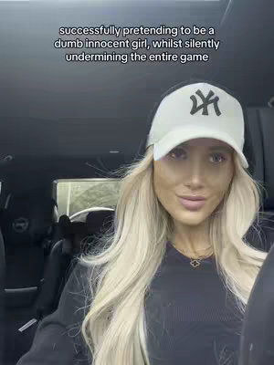 A woman with long blonde hair is seated inside a vehicle. She wears a white baseball cap with a black logo, a black top, and a gold necklace. The background includes car interior and a glimpse of the outside.