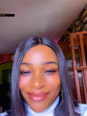 A selfie-style shot featuring a woman from a slightly elevated angle. She has dark hair parted in the middle, cascading around her face. Her gaze is directed downward with eyelids partially closed. The focus is on her face, displaying smooth, illuminated skin with pronounced, glossy lips. She wears a white turtleneck, set against an out-of-focus background including visible interior elements like a door frame.