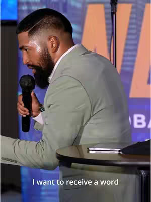 A man with a trimmed beard and short haircut is speaking into a microphone on a stage. The background is a cityscape with the word 'GLOBAL' partially visible. He is wearing a light green suit and is standing behind a dark table. At the bottom of the image, the text 'I want to receive a word' is overlaid.