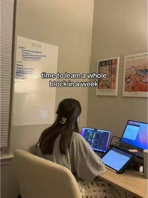 A person is sitting at a desk with two laptops and a tablet; the person is facing away, revealing their head and back as they look at the screens, with a white board and framed pictures in the background.