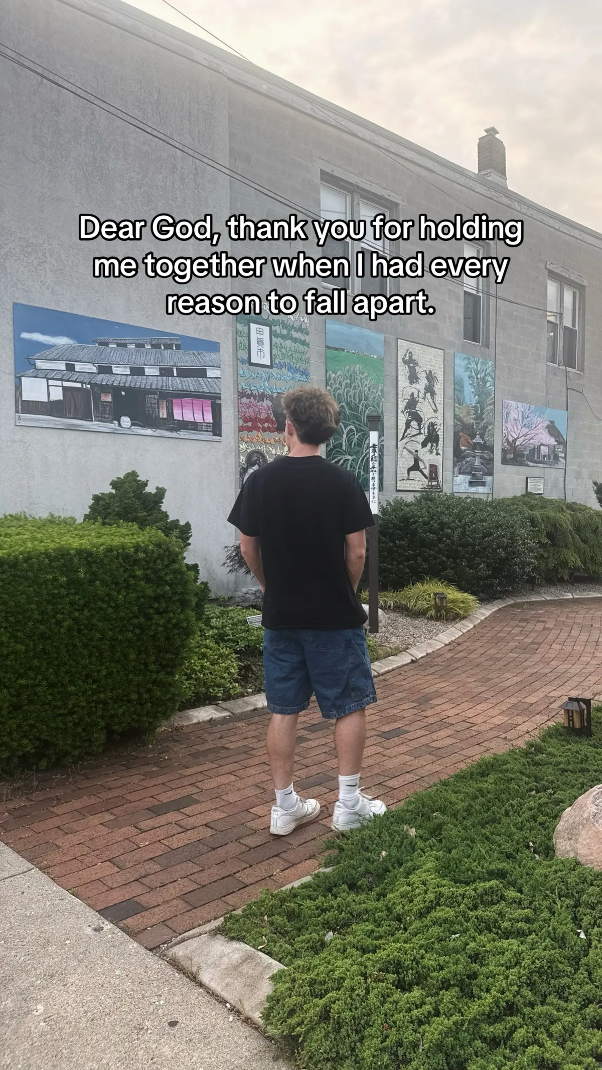 A man stands with his back to the camera, facing a building with murals. Text overlayed on the image. The scene is outdoors on a cloudy day.