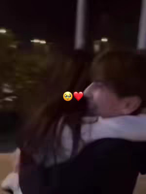 An out-of-focus image featuring a close embrace between two individuals at night. The primary colors are dark, offset by the glowing lights from nearby structures.