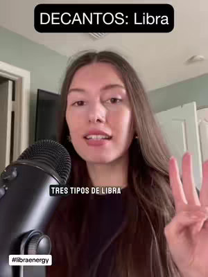 A brown-haired woman with a neutral expression is speaking in front of a grey microphone. The text overlay shows the phrase "DECANTOOS: Libra" in a black rectangular box.