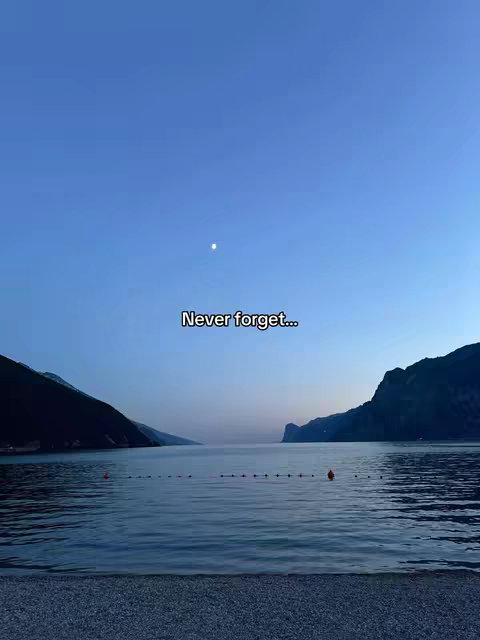 The image shows a scenic landscape at dusk. The sky is a gradient of deep blue to lighter blue, with a bright moon visible. Silhouetted mountains are on either side of a calm body of water. A row of buoys stretches across the water's surface. The foreground is a pebble beach.