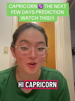 A woman with glasses is centered in the frame, wearing a green shirt, appears to be speaking directly to the camera. Overlaid are two text boxes in neon green and red, which says 'CAPRICORN ♑️ THE NEXT FEW DAYS PREDICTION WATCH THIS!!!' and 'HI CAPRICORN'. The background appears to be an interior, with a door and light switch visible.