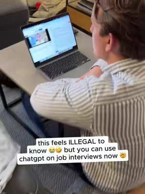 A man is seated at a desk with a laptop, with the screen showing a video call. The man is angled to the right, with his back to the camera, wearing a striped shirt. A text overlay at the bottom states, "this feels ILLEGAL to know 🤭 but you can use chatgpt on job interviews now 🤭". The setting appears to be a home office, with a gray couch and a small desk in view. The lighting seems natural.