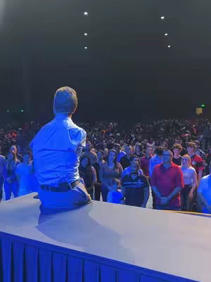 A man with dark hair faces away from the camera, kneeling on a blue surface. He appears to be speaking to a large crowd of people, who are facing him. The lighting is dramatic, with blue and cool tones illuminating the scene.