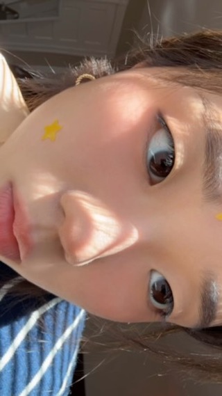 A close-up shot of a person's face, angled from the side. The face is well-lit, with soft shadows. The person has prominent eyes and a slight smile, with a small star-shaped sticker on one cheek. The background is softly blurred, suggesting an indoor setting.