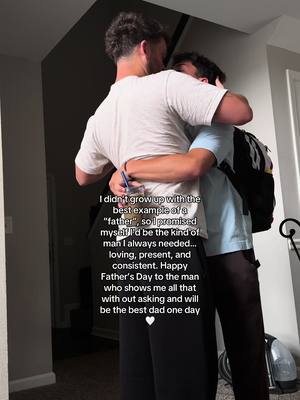 Two men are embracing in a close hug, viewed from the shoulders up. The man on the left, whose back is towards the camera, appears to be wearing a white t-shirt. The man on the right is partially visible, wearing a light blue shirt and a backpack. Text overlay is included on the image.