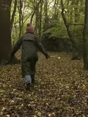 A person walks away from the camera into a forest filled with fallen leaves. The person wears a dark coat and a pink beanie.