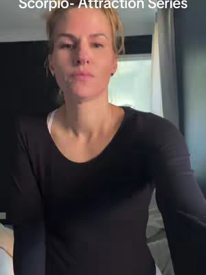 A medium shot of a fair-skinned woman with blonde hair, looking at the camera with a neutral expression. She's wearing a black long-sleeved top and the background is a blend of a dark wall and a window with a curtain.